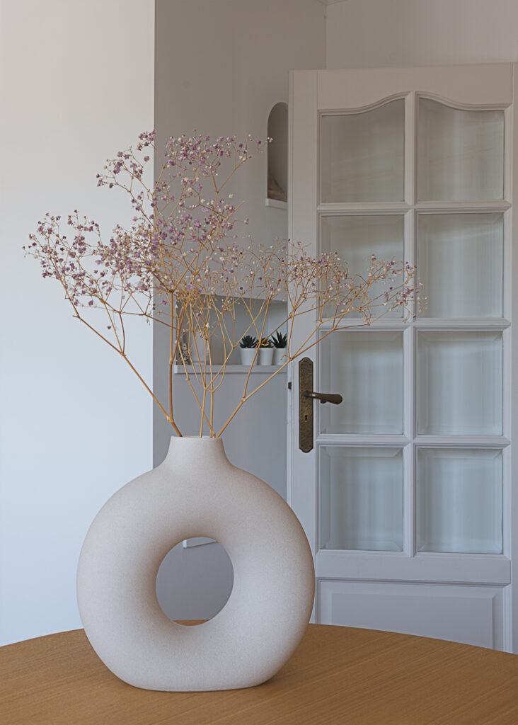 Détails d'une photographie immobilière. Un vase rempli de brindille fleuries est posée sur une table en bois. Au fond, il y a une porte blanche vitrée