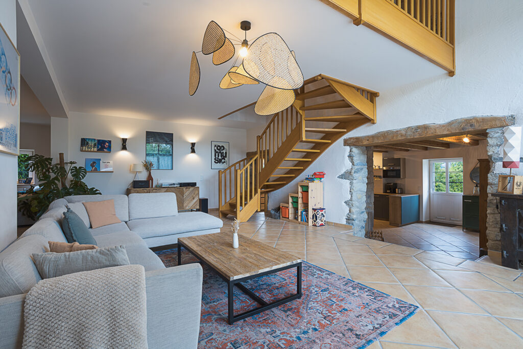 Photographie immobilière en Haute-Savoie. Un grand Salon surélevée dans cette ferme réhabilité . un escalier en bois donne à l'étage avec une mezzanine. et une grande ouverture avec un linteau en bois mène à la cuisine.